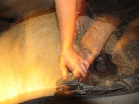 1978 Ford Bronco - A hand is shown reaching under the carpeted interior of a vehicle, revealing a rusty underbody.
