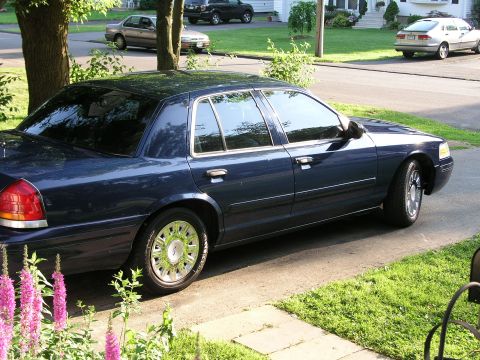 1999 Ford Crown Victoria - A dark blue sedan is parked on a driveway, with other vehicles and houses in the background.