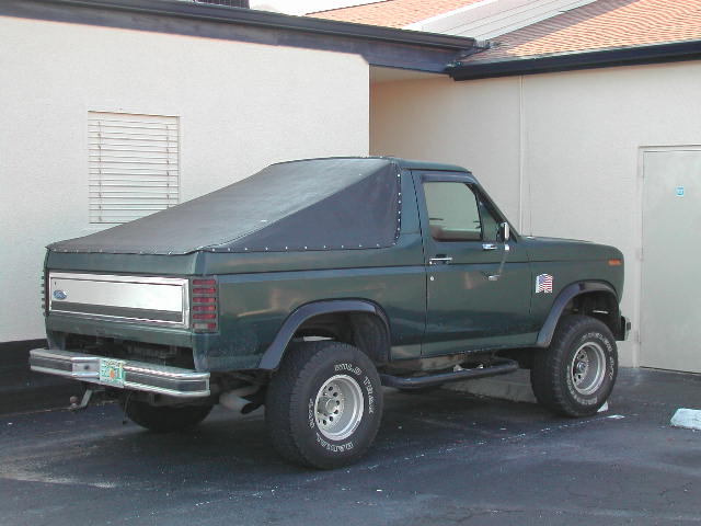 Dark green lifted Ford Bronco with black soft top and large tires, rear three-quarter view beside a tan building