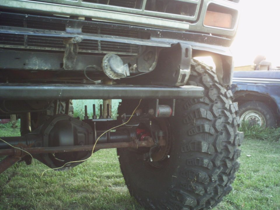 Front suspension and large off-road tire of a modified truck; another vehicle is visible in the background