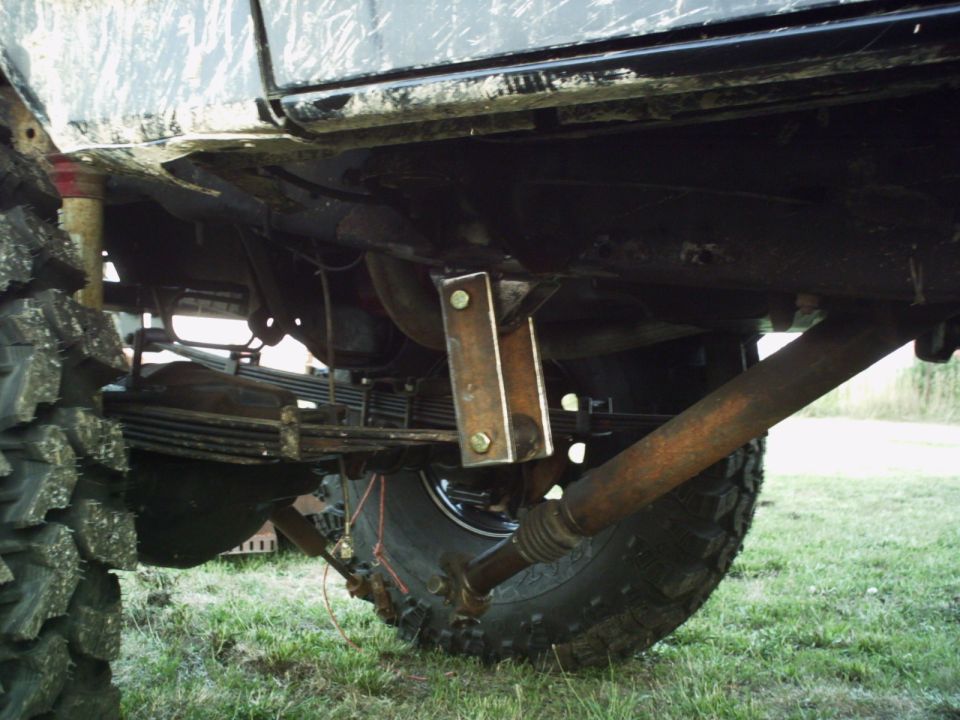 Underside view of a vehicle with large tires, showing leaf springs, drive shaft, and custom metal brackets bolted to the frame