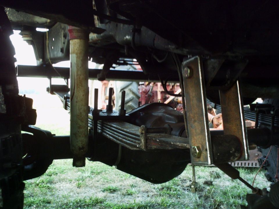 Undercarriage of a vehicle showing leaf springs, a shock absorber, and custom metal brackets. A tractor is visible in the background