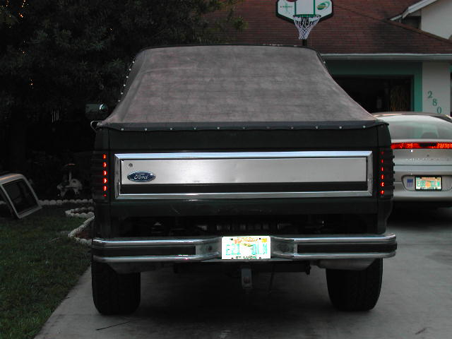Rear view of dark green Ford with fabric top cover, aluminum tailgate panel, and chrome bumper in a driveway at dusk