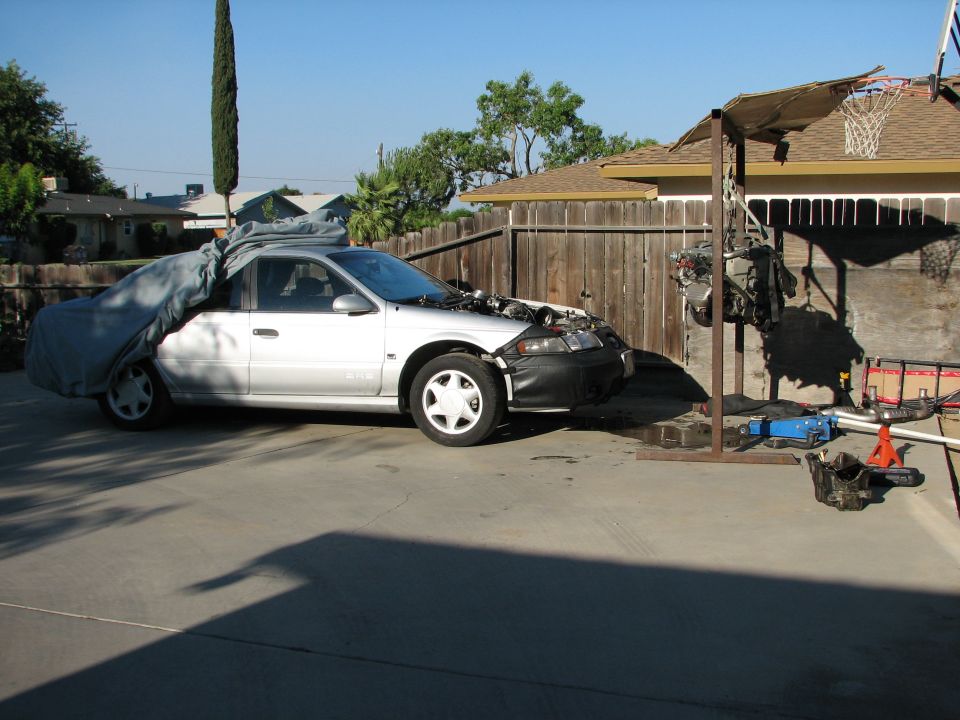 Silver sedan with car cover on rear half, hood open, engine removed and hanging from a stand. Some tools on the ground
