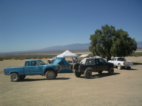 1985 Ford Ranger - Several off-road trucks are parked in a desert area with tents set up, under a clear blue sky.