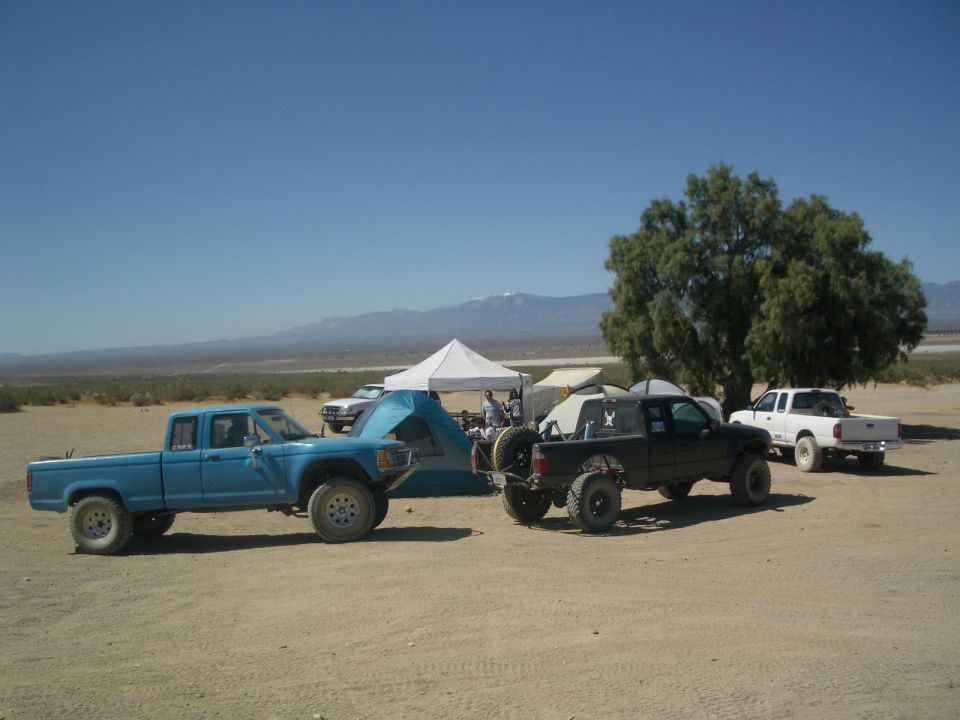 Blue and black off-road trucks parked near tents in a desert landscape with mountains in the background