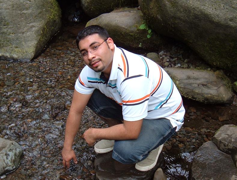 Man in striped shirt and jeans crouching by a rocky stream, with a SUPERMOTORS watermark