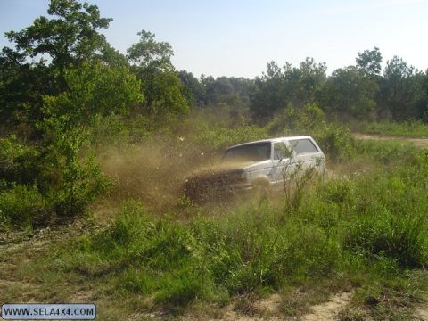 1995 Ford Bronco - A white SUV navigates a muddy, overgrown trail, sending dirt flying.
