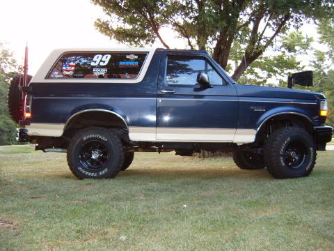 1993 Ford Bronco - A dark blue and tan Ford Bronco is shown from the side, parked on a grassy area with trees in the background.