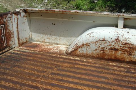 1963 Ford F-250 - The interior of a heavily rusted truck bed shows significant wear and tear on its ribbed flooring and white wheel well.