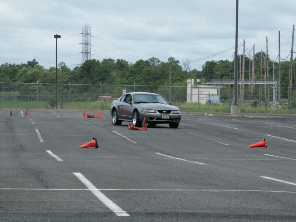 Silver Ford Mustang coupe with a license plate visible, driving on a paved lot with orange cones