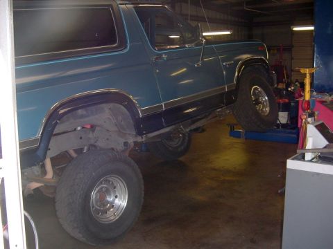 1984 Ford Bronco - A teal Ford Bronco is elevated on a lift inside a garage, revealing its undercarriage and suspension system.