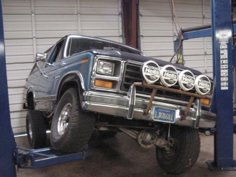 1984 Ford Bronco - Two-tone blue and silver 1980s Ford full-size SUV on a blue two-post shop lift, 3/4 front view, with the front axle drooped to show suspens…