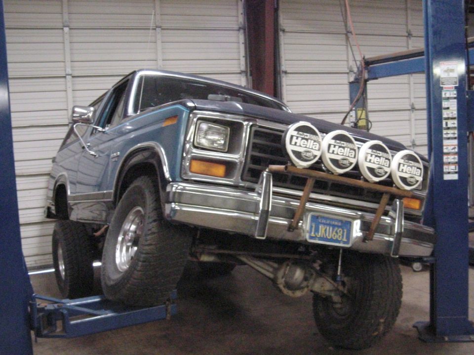 Blue two-tone 1980s Ford SUV on a shop lift with drooped front axle and four Hella lights