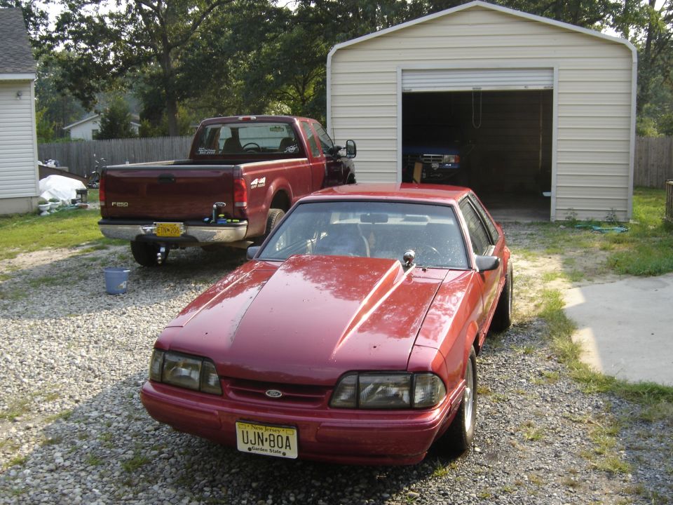 Red coupe with aftermarket hood scoop and UJN-80A license plate parked in gravel driveway. Burgundy pickup truck and garage in background