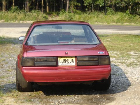 1988 Ford Bronco - Rear view of a red coupe with a "MUSTANG" badge, indicating it is likely a Ford Mustang from the late 1980s or early 1990s.