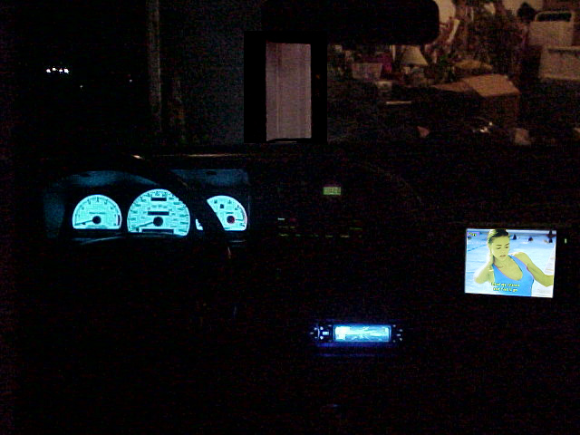 Dark car interior at night with glowing blue-green gauges, blue stereo display, and small video screen