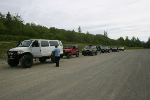 2003 Jeep TJ - Several off-road vehicles are parked in a line on a gravel lot with a wooded hillside behind them.