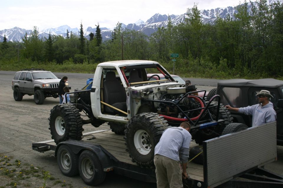 Custom pickup truck with huge tires on a trailer, another Jeep and a dark SUV nearby, mountains in background