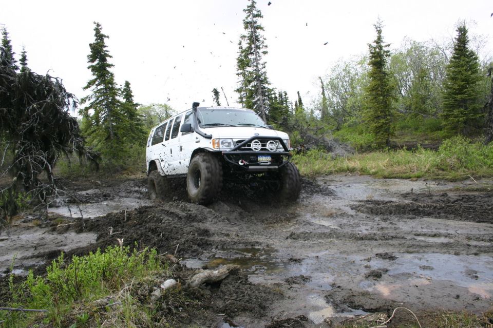White SUV with snorkel and aftermarket bumper driving through a muddy trail with trees in the background