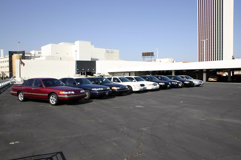 Row of Crown Victoria sedans in a parking lot, dark red car in front, buildings and flag behind