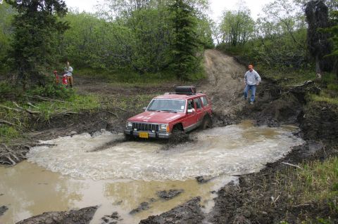 2003 Jeep TJ - A red Jeep Cherokee XJ navigates a challenging muddy water crossing on an off-road trail.