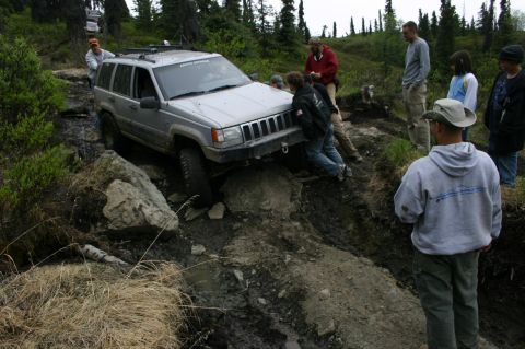 2003 Jeep TJ - A group of people watch as a silver Jeep Grand Cherokee navigates a challenging, muddy off-road course.