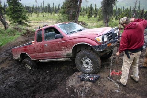 2003 Jeep TJ - A red pickup truck is stuck in deep mud while a person operates a winch.