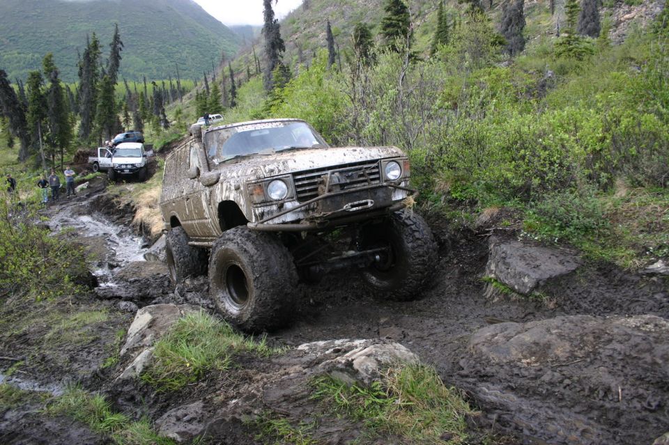 Muddy SUV navigating a rocky, wet trail with trees and mountains in the background. Another white SUV is visible further up the path
