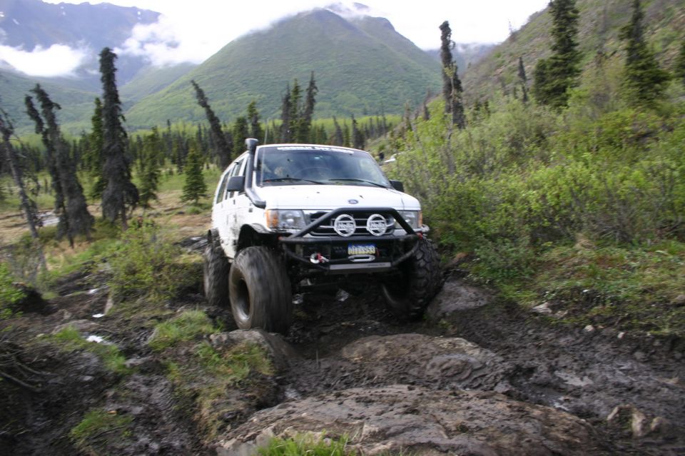 White SUV with snorkel and off-road bumper navigating a muddy, rocky trail with mountains in the background