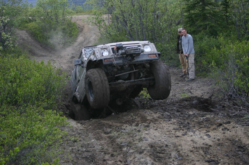 Off-road vehicle with large tires struggling in a muddy ditch on a dirt trail, two people watching