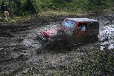2003 Jeep TJ - A red 2003 Jeep TJ Wrangler navigates a muddy trail, kicking up water and mud as it gets stuck.