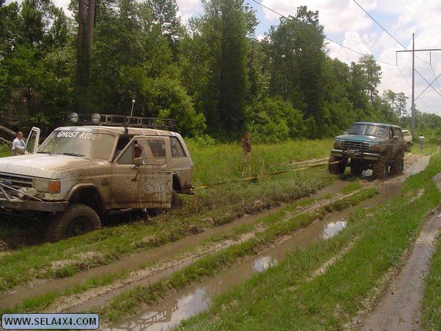 Muddy green SUV pulling a white SUV with "GHOST RIDER" on windshield through a muddy track