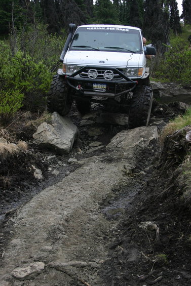 2000 Ford E350 - A white Ford van or truck with a lifted suspension and large off-road mud tires is shown from the front, crawling over a rocky, muddy trail…