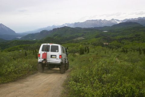 2000 Ford E350 - Rear view of a white lifted 4x4 van on oversized tires driving away up a dirt track through green brush, with a red fuel can and Hi-Lift ja…