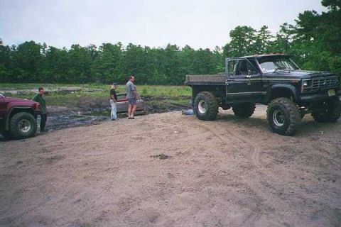 1990 Ford Bronco - A black pickup truck with oversized tires sits on a dirt lot next to a maroon SUV and several people.