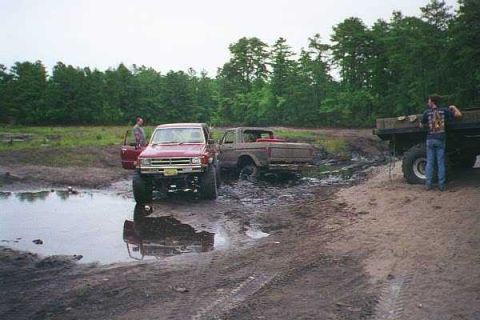 1990 Ford Bronco - Off-road trucks navigate a muddy terrain with onlookers present.