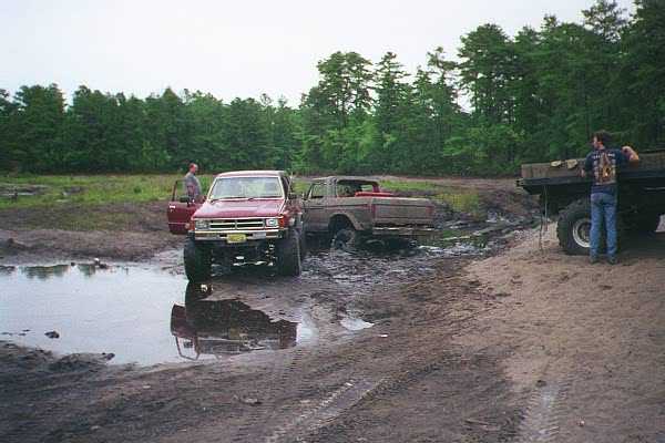 Two off-road trucks stuck in mud, one red and one brown, with people standing nearby