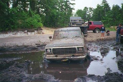 1990 Ford Bronco - This muddy scene shows a classic Ford F-Series truck deeply stuck in a mud pit, with other vehicles and people nearby.