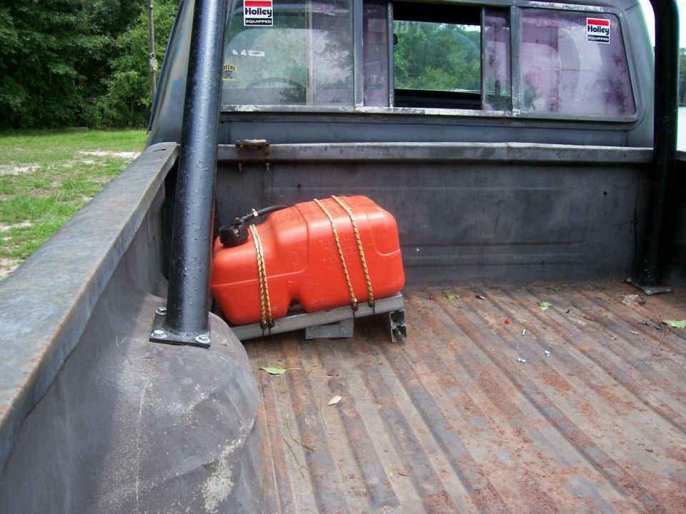 Truck bed interior with orange auxiliary fuel tank secured by ropes. Holley stickers visible on rear window