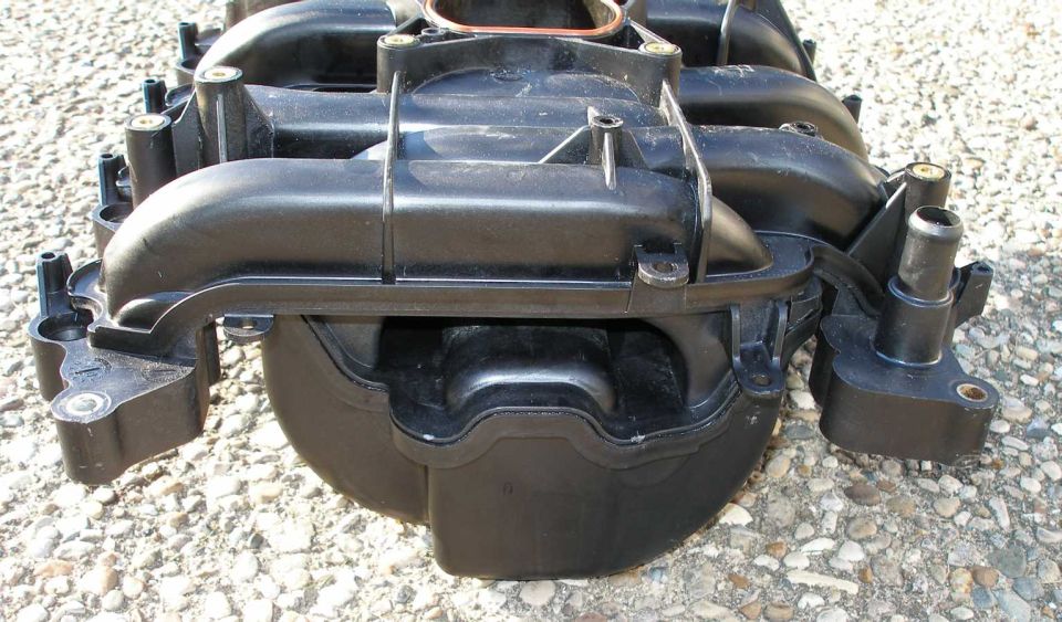 Rear of a black composite V8 intake manifold sitting on gravel