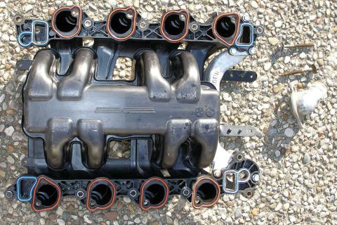 1998 Ford Crown Victoria - A disassembled engine intake manifold lies on a gravel surface, with various bolts and a metal fitting nearby.