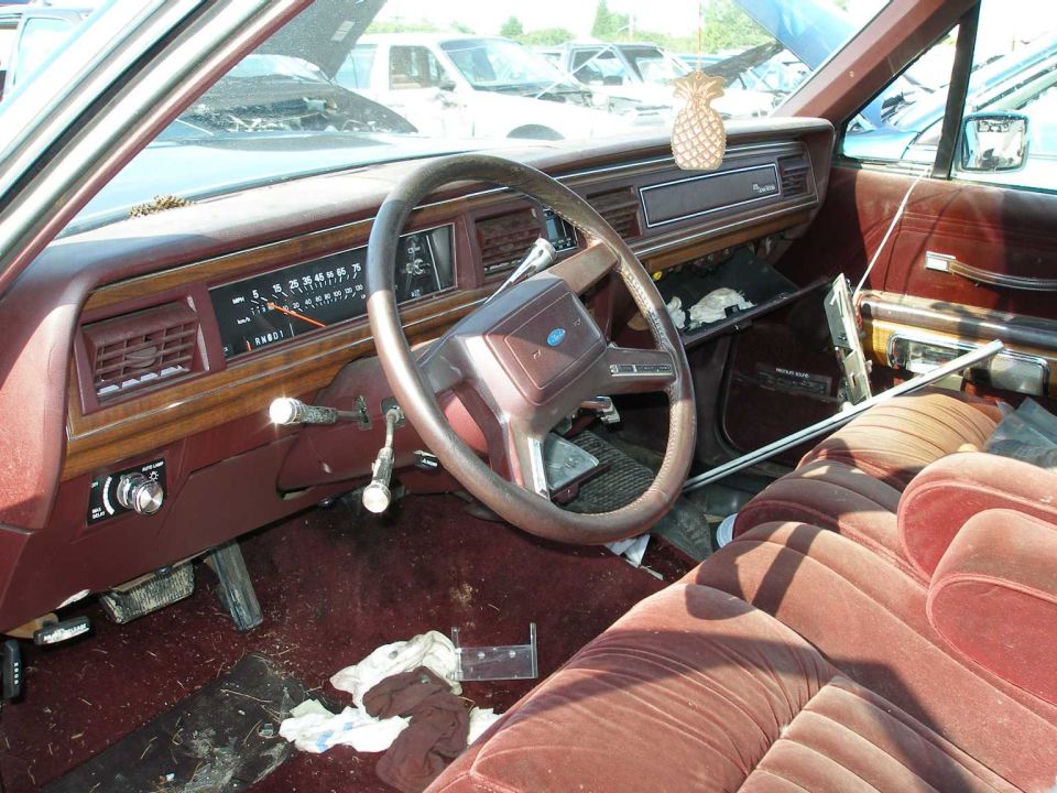 Burgundy interior of an old Ford sedan with red velour seats and Ford steering wheel in a junkyard