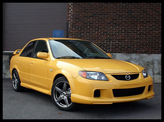 Yellow sedan parked in front of a brick building. It has a spoiler and aftermarket wheels