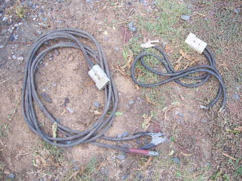 1983 Ford Bronco - Jumper cables lie on the ground, one set coiled and the other loosely arranged.