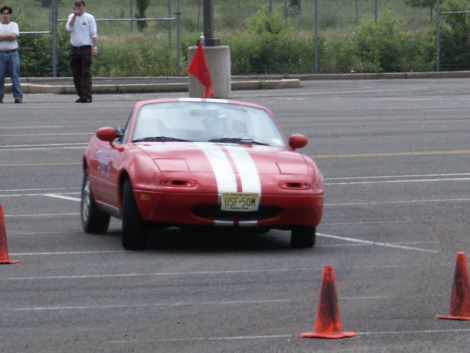 Red convertible sports car with white racing stripes and license plate USF-50M on a paved lot with orange cones