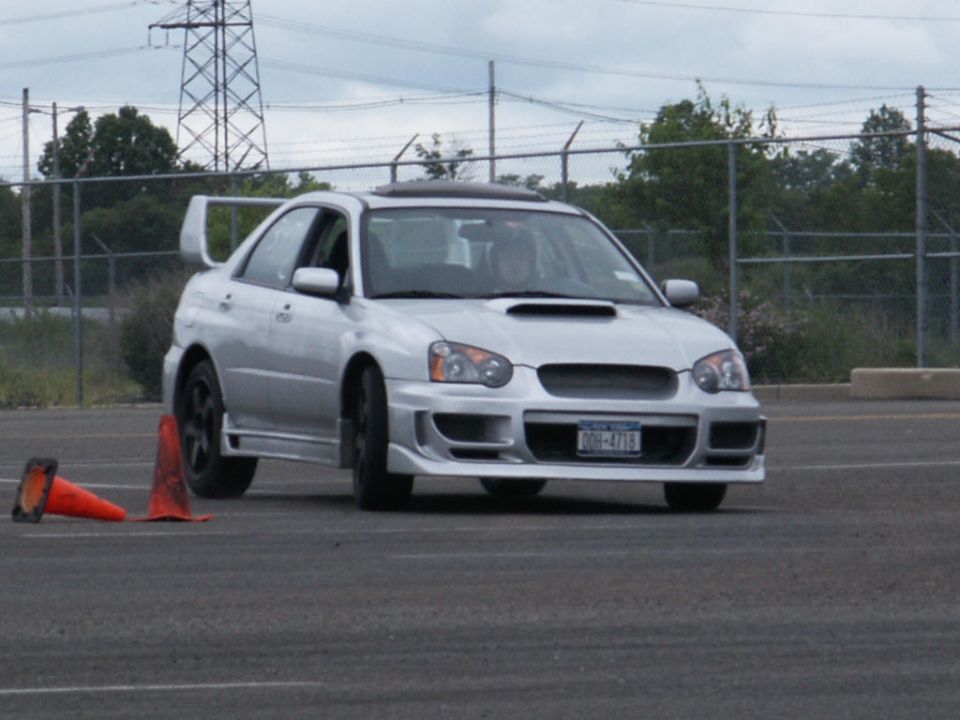 Silver Subaru Impreza WRX with a rear spoiler and aftermarket body kit, front view, on asphalt with orange cones