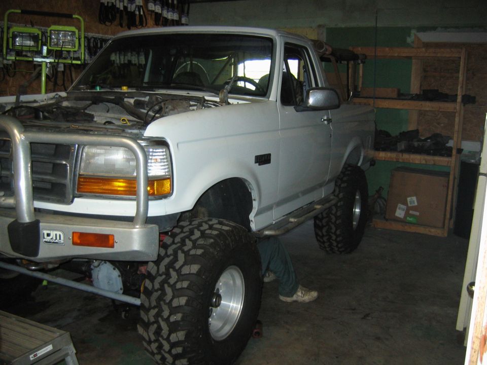 White lifted Ford Bronco on mud tires in a garage, hood open, front-left view
