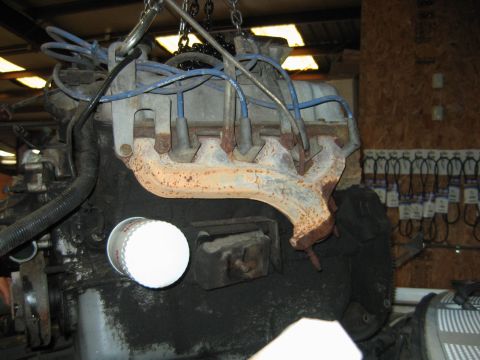 1992 Ford Bronco - An engine block suspended by chains, showing a rusty cast-iron exhaust manifold, blue spark plug wires, and an oil filler cap, with an inta…