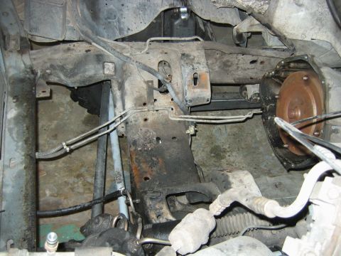 1992 Ford Bronco - Underbody view of a vehicle chassis showing the front frame crossmember, rusty steel framerails, brake/fuel lines, and a bell housing/flywh…